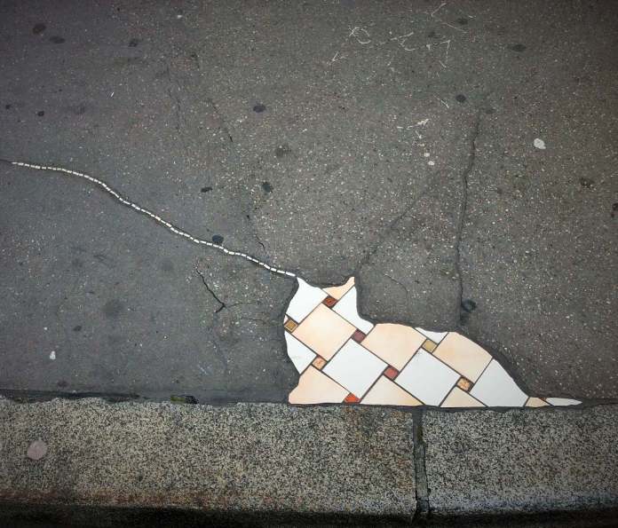 A playful mosaic by Ememem featuring a cat shape created from beige and orange tiles, integrated seamlessly into the pavement. The design uses a crack in the sidewalk to represent the cat’s tail, extending into the surrounding area. This whimsical artwork transforms urban damage into a charming and creative scene, inviting viewers to see beauty in imperfection.