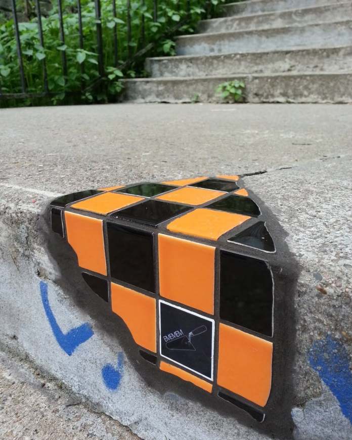 A mosaic by Ememem embedded in a cracked corner of a concrete stair. The design features a bold checkerboard pattern in orange and black tiles, accented with a signature tile displaying Ememem’s logo. This small but striking piece highlights the artist's ability to transform minor urban damage into playful and vibrant street art.