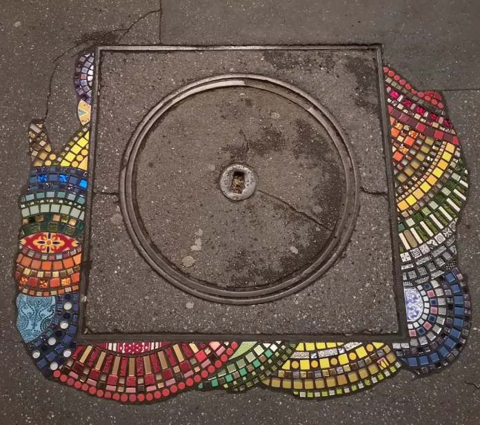 A vibrant mosaic by Ememem integrated around a circular manhole cover. The design features a colorful rainbow-like pattern of tiles in red, yellow, green, and blue, flowing outward from the cover and blending seamlessly with the surrounding pavement. This artwork transforms the cracked and worn manhole area into a striking piece of urban art, celebrating creativity in unexpected places.