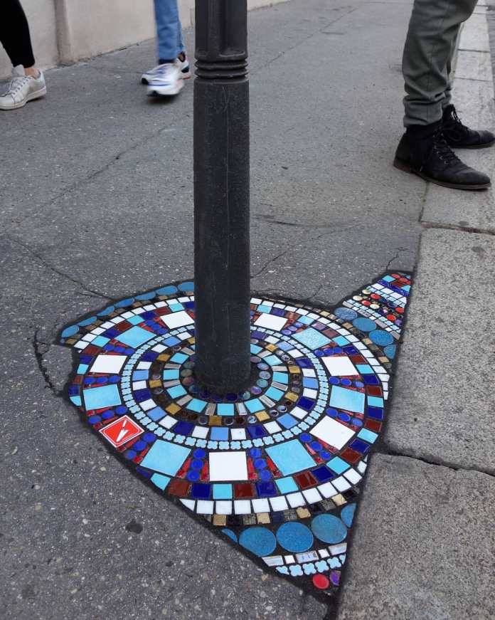A vibrant mosaic artwork by Ememem in Lyon, France, surrounding the base of a black streetlamp. The intricate design features geometric patterns in blue, white, and red tiles, meticulously arranged to repair a crack in the pavement. Pedestrians' legs in the background hint at the urban setting, emphasizing how this art transforms everyday spaces into striking visual experiences.