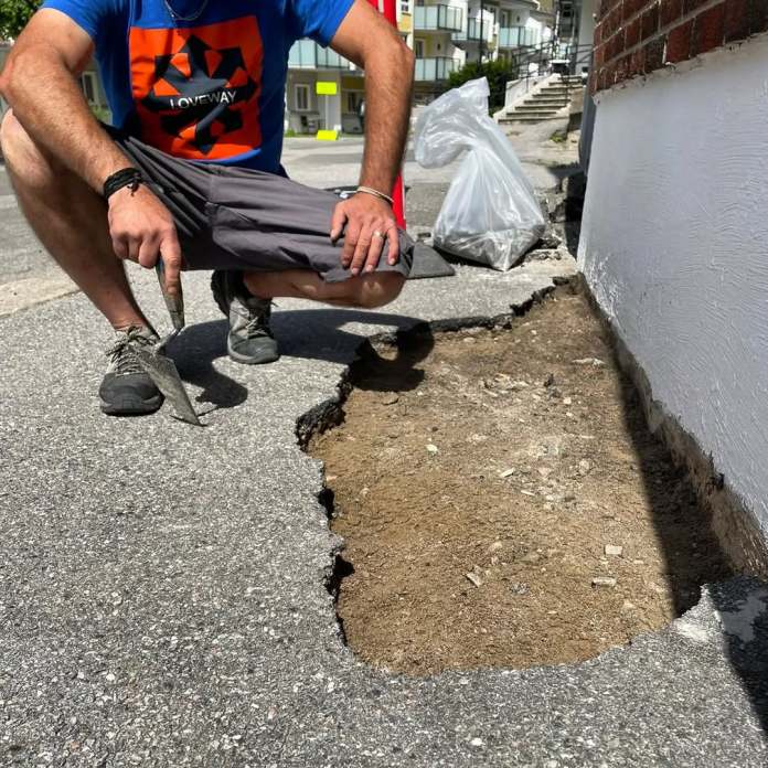 An image of Ememem preparing a damaged section of pavement for one of his mosaic installations. The artist, dressed in a blue shirt with a bold graphic design, kneels next to a large pothole filled with dirt, holding a trowel. The scene captures the beginning stage of Ememem’s creative process, where urban damage is about to be transformed into vibrant mosaic art.