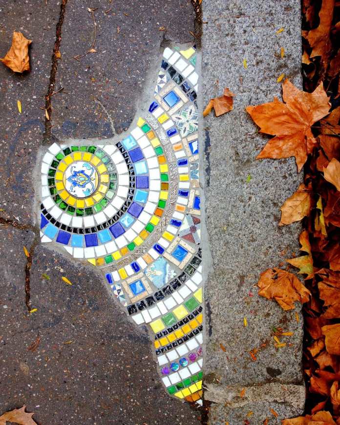 A mosaic by Ememem integrated into a cracked sidewalk, featuring vibrant patterns in green, blue, yellow, and white tiles. The circular design starts with a yellow centerpiece and radiates outward with intricate geometric details, blending seamlessly into the pavement. Autumn leaves scattered around the edge add a seasonal touch, contrasting the colorful artwork with the natural environment.