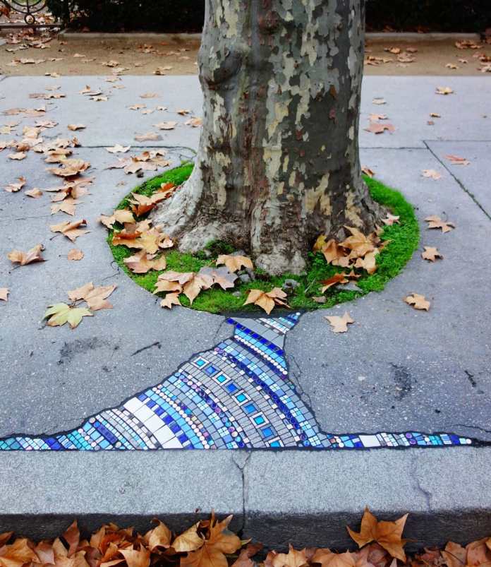 A mosaic by Ememem surrounding the base of a tree in a cracked sidewalk. The design features intricate patterns in blue, white, and purple tiles, radiating upward like a tree trunk's extension. Green moss and autumn leaves frame the artwork, blending natural and creative elements to transform an otherwise damaged urban area into a visually captivating scene.