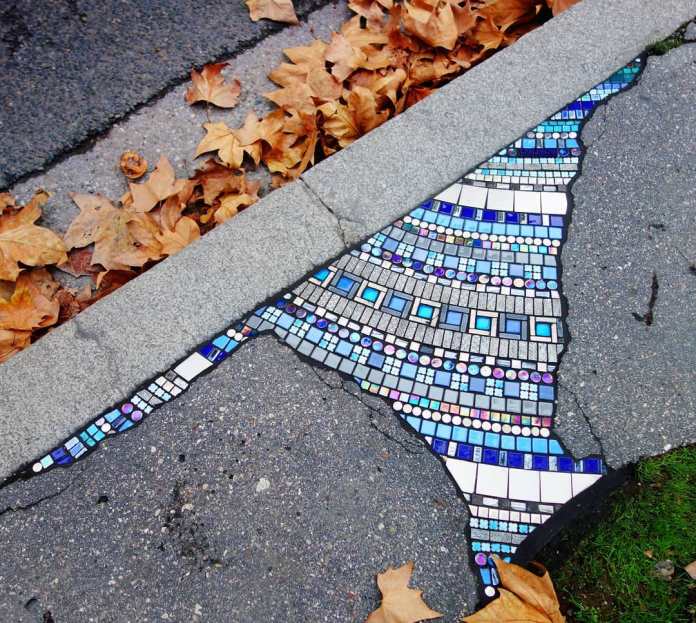 A close-up of a mosaic by Ememem, seamlessly integrated into a cracked sidewalk. The artwork features intricate geometric patterns in shades of blue, white, and purple tiles, radiating in a flowing design that follows the contours of the crack. Autumn leaves scattered along the edge add warmth and contrast, emphasizing the harmony between art and nature.