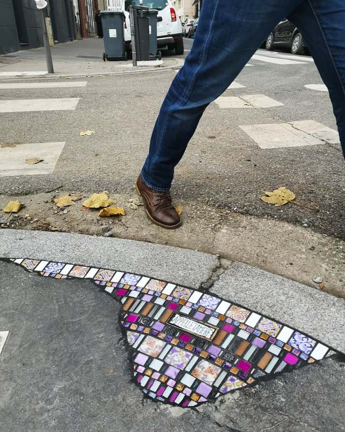 A mosaic artwork by Ememem seamlessly integrated into a cracked curb. The design features a mix of intricate tiles in purple, white, and orange hues, complemented by patterned elements that add depth and character. A passerby steps over the mosaic, emphasizing its placement in a functional urban space. Fallen autumn leaves frame the scene, creating a balance between artistry and the natural environment.