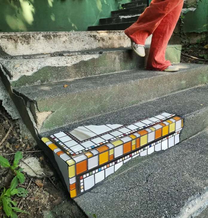 A mosaic by Ememem integrated into a damaged concrete stair corner. The artwork features geometric patterns in warm shades of orange, yellow, and white tiles, forming a striking visual contrast against the worn gray steps. A person in vibrant red pants walks up the stairs, adding a dynamic human element to this creative urban repair.