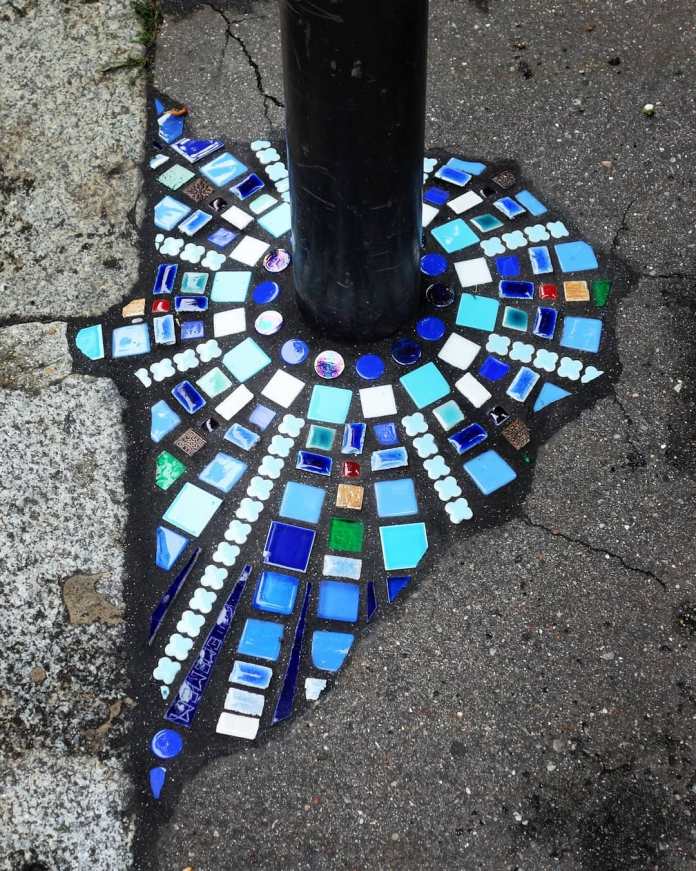 A mosaic by Ememem encircling the base of a black pole, featuring a vibrant geometric design in shades of blue, white, and green tiles. The mosaic transforms a cracked section of pavement into a radiant artwork, showcasing intricate patterns that radiate outward. This piece exemplifies Ememem's talent for turning urban imperfections into beautiful street art.