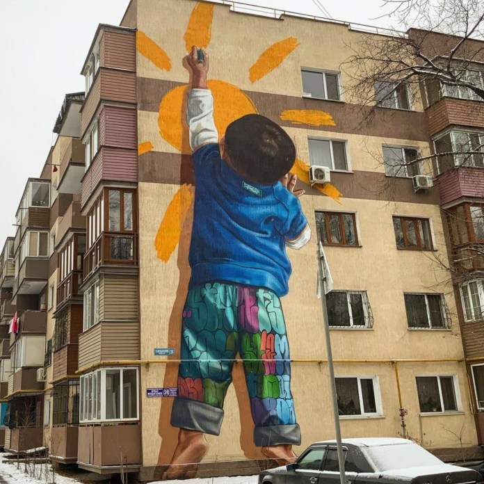 Large mural of a child drawing a yellow sun on an apartment building in Almaty, Kazakhstan. The child wears colorful pants and a blue hoodie, painted in a realistic style with upward perspective by artists TANAI and Ali Zakir.