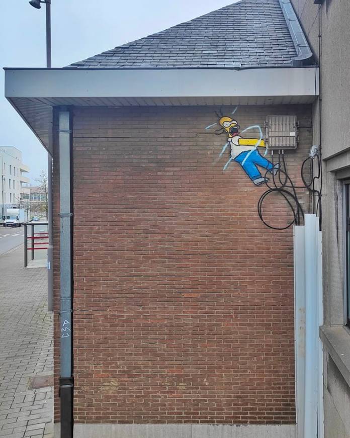 Street art by Oakoak showing Homer Simpson being electrocuted by a real electrical box. His body appears shocked and twisted, integrated seamlessly into the building’s wiring.