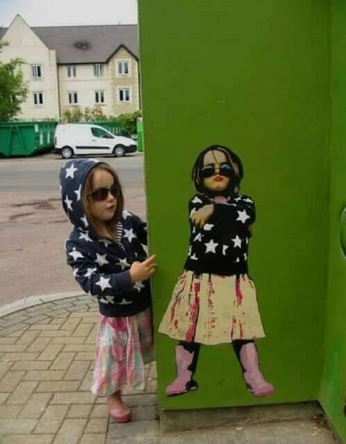 Real child mimics a street art portrait of herself wearing the same starry hoodie and sunglasses. Painted on a green utility box by Andy Dice Davies.