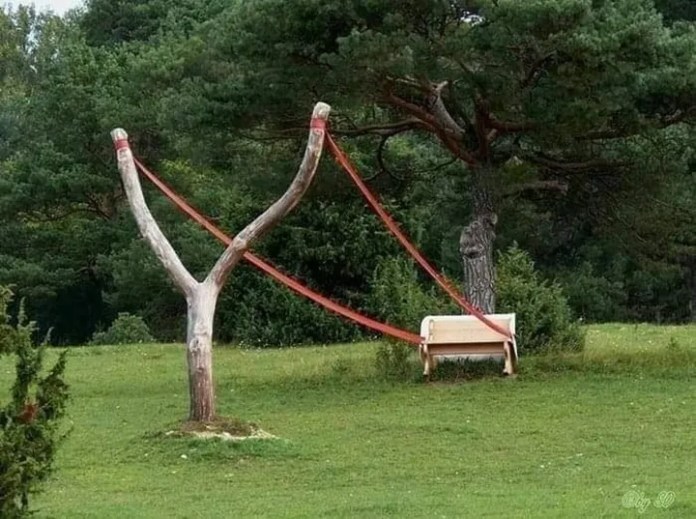 Outdoor sculpture in a grassy park in Germany showing a bench slung between two red straps attached to a large Y-shaped tree branch, designed to resemble a giant slingshot.