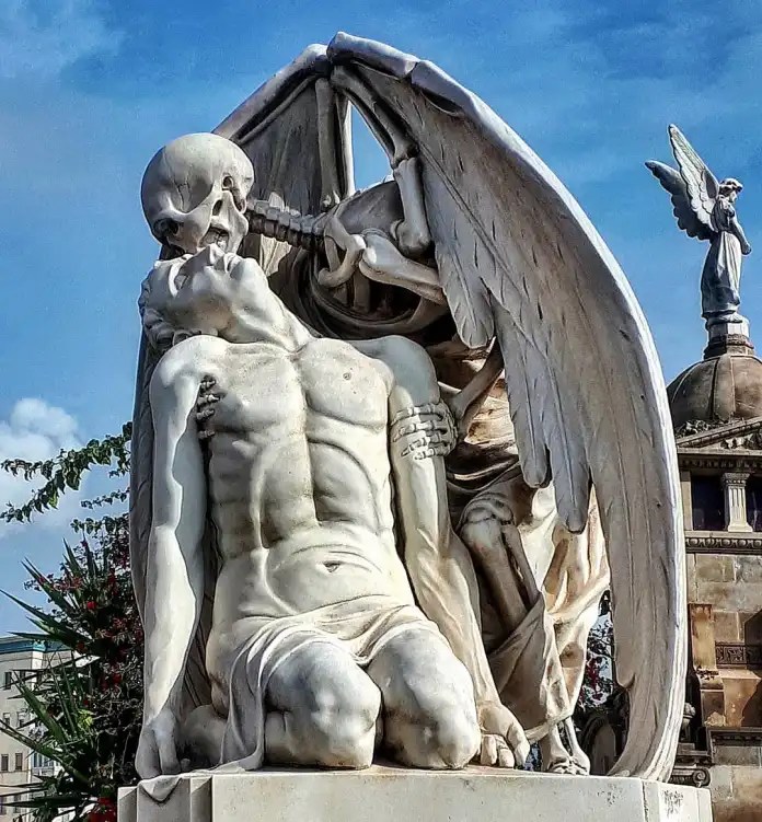 Marble statue in Barcelona’s Poblenou Cemetery depicting a winged skeleton embracing and kissing the forehead of a limp male figure kneeling on a plinth. Background includes other cemetery sculptures.
