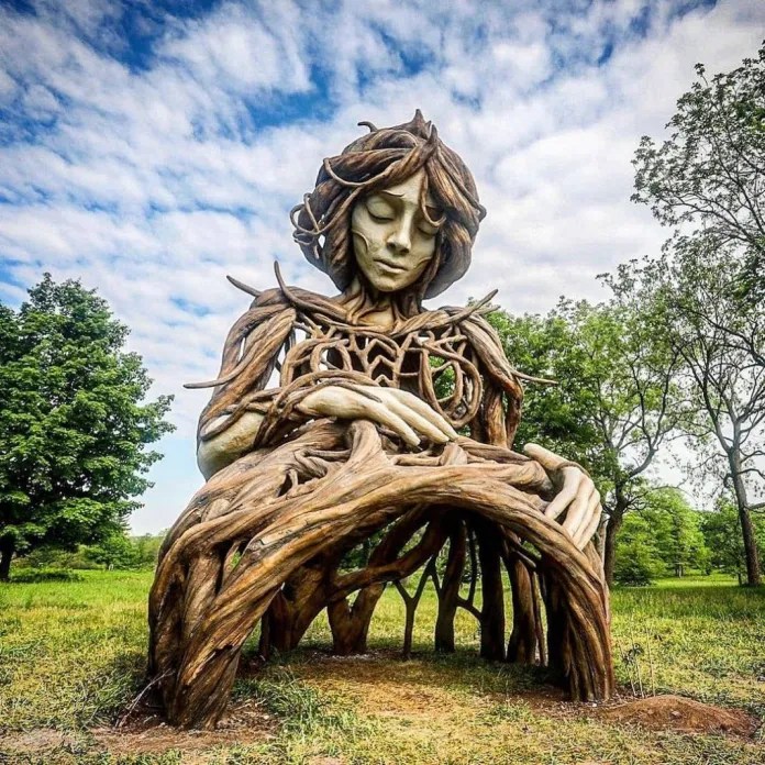 Giant wooden sculpture by Daniel Popper at The Morton Arboretum in Illinois showing a serene woman composed of interwoven tree roots and branches, set in a green meadow under a blue sky.