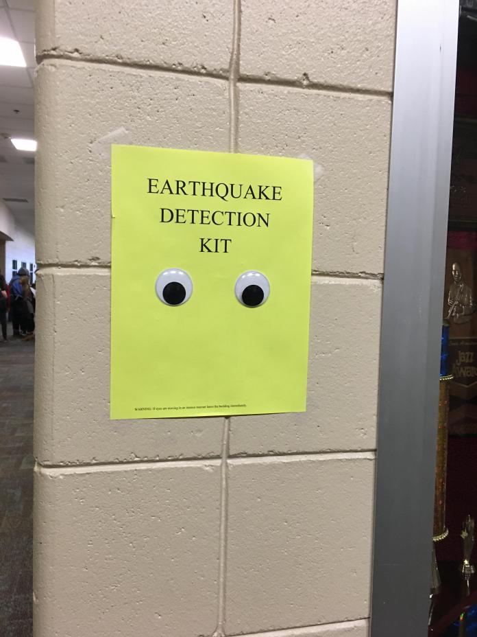 A bright yellow paper labeled "EARTHQUAKE DETECTION KIT" taped to a hallway wall with two googly eyes attached below the text.
