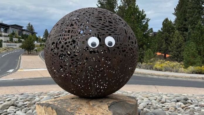 A brown perforated metal sphere sculpture on a rock pedestal, with googly eyes added to the front, placed near a road in Bend, Oregon.
