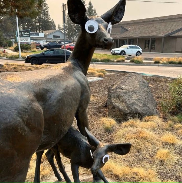 Bronze sculpture of a doe and fawn in a landscaped roundabout in Bend, Oregon, both fitted with oversized googly eyes, creating a humorous contrast with their naturalistic forms.