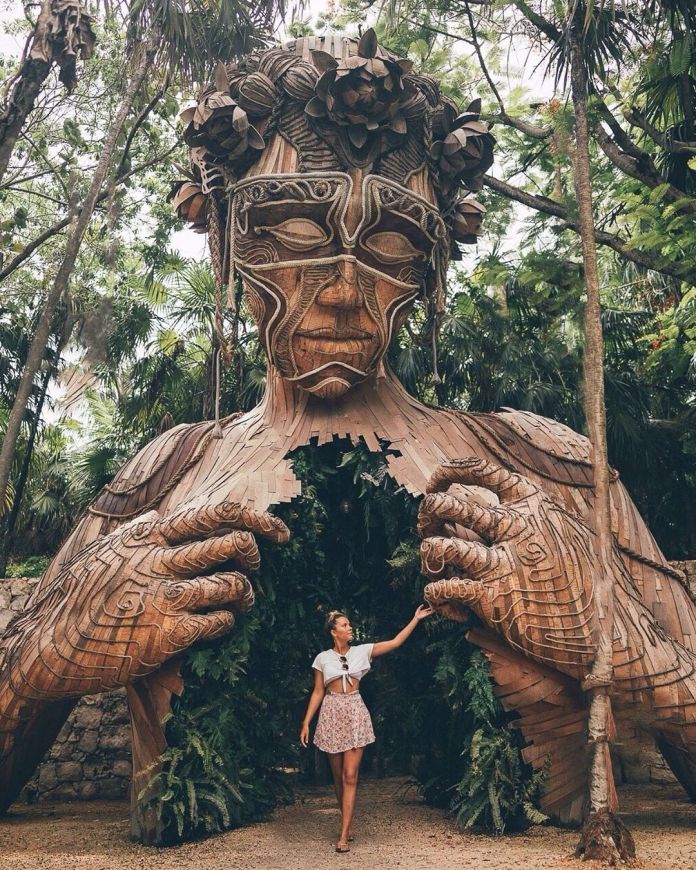 Towering wooden sculpture of a person with hands opening their chest, revealing a path surrounded by plants, located in a forested area in Tulum, Mexico.