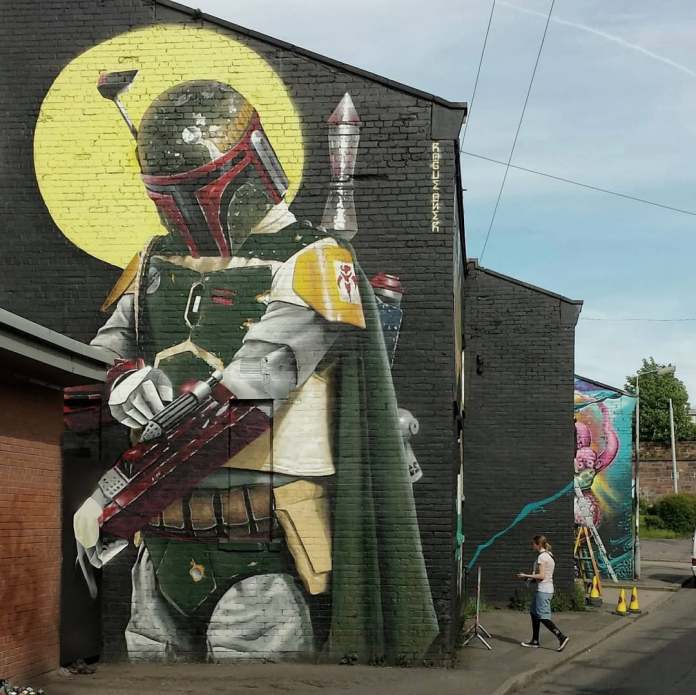 A mural by Bobby Rogue-One in Glasgow, Scotland, created as a tribute to the late Jeremy Bulloch, who portrayed the iconic Star Wars character Boba Fett. The artwork, painted on a brick wall at Yardworks, features a highly detailed depiction of Boba Fett in his green and gray Mandalorian armor, complete with a cape, blaster, and jetpack. A glowing yellow circle behind his helmet gives the piece a striking, almost halo-like effect. The mural captures the stoic essence of the character and celebrates the legacy of both the actor and the Star Wars universe. A passerby is visible near the artwork, adding a sense of scale to the mural.