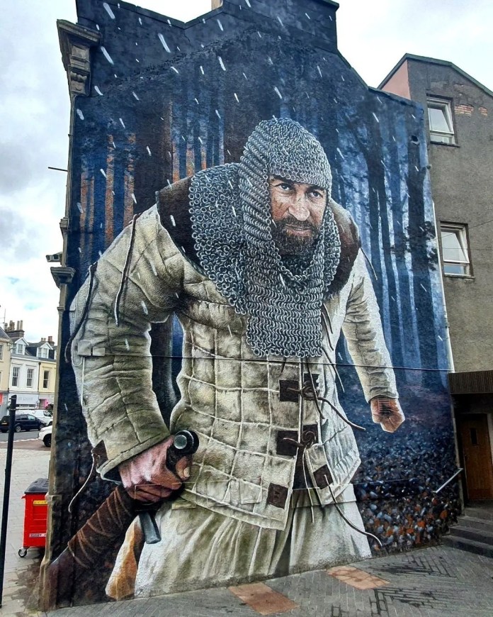 A mural by Bobby Rogue-One in Lanark, United Kingdom, depicting the historical figure William Wallace. The artwork shows Wallace in chainmail and padded armor, gripping the hilt of his sword as he flees through Clyde Forest. Snow falls around him, adding a dramatic touch to the scene, which commemorates the aftermath of Wallace sacking and burning the castle in Lanark. The forest backdrop, painted in deep blues and browns, enhances the tension and historical atmosphere. This is the second of two William Wallace murals in Lanark, created as part of the town’s Wallace public space, a tribute to the Scottish hero.