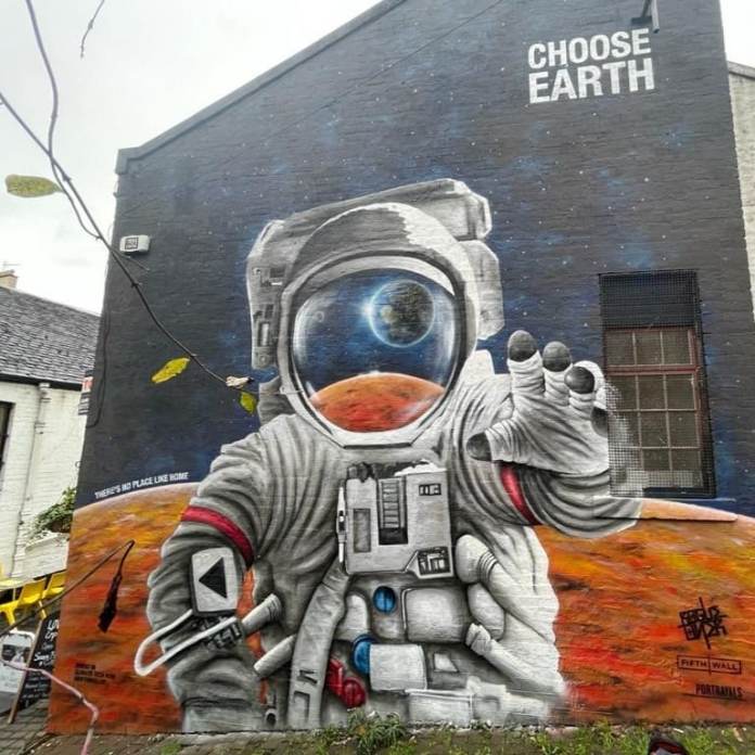 A mural by Bobby Rogue-One in Cresswell Lane, Glasgow, created in collaboration with The Portrayals and commissioned by Fifth Wall VC for COP26. The artwork features a hyper-realistic astronaut standing on the surface of Mars, reaching out towards Earth, which is beautifully reflected in the visor of the astronaut’s helmet. Above the mural, the bold text reads, 'CHOOSE EARTH,' emphasizing the message of prioritizing climate action. The lower text states, 'There’s no place like home,' reinforcing the call to focus resources on saving the planet. The mural was painted using a combination of carbon-absorbing paint for the larger areas and acrylic and water-based spray paints for intricate details. The background showcases a cosmic scene with stars and Mars’ surface, encapsulating the theme of longing and urgency to protect Earth.