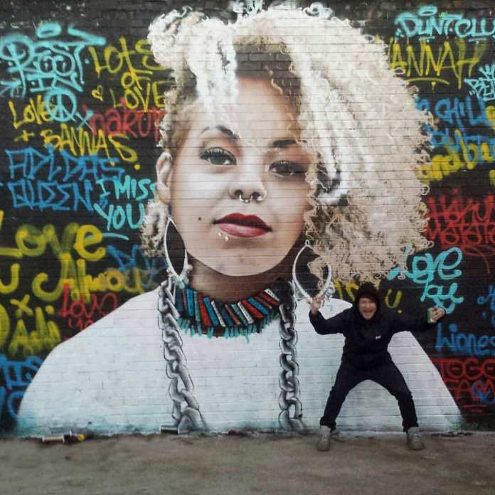 A mural by Bobby Rogue-One in Glasgow, created as a heartfelt tribute to Hannah, a beloved friend of the artist’s acquaintance, Wee Rab. The portrait depicts Hannah with striking blonde curls, a nose ring, and colorful beaded jewelry, set against a vibrant graffiti-style background filled with messages and dedications from her friends. The messages, written in various colors, add a deeply personal touch to the piece. Wee Rab stands in front of the mural, smiling proudly, reflecting the emotional connection shared by all who participated in this tribute. The mural radiates warmth and community spirit, honoring Hannah's memory.