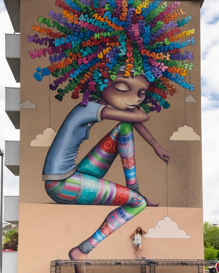 Large mural of a young girl with patterned leggings sitting with eyes closed, her oversized hair filled with multicolored words such as “diversity” and “égalité,” painted by Vinie in Chatou, France.