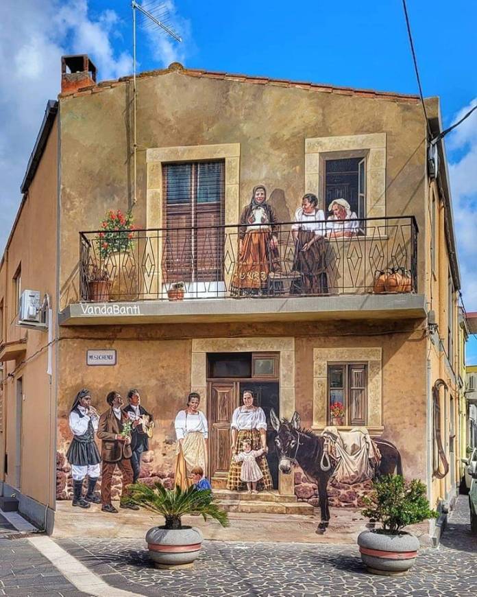 Trompe-l’oeil mural of villagers in Sardinia wearing traditional clothing, with a donkey and people gathered outside a rustic house, painted by Vanda Banti in Zeddiani, Italy.