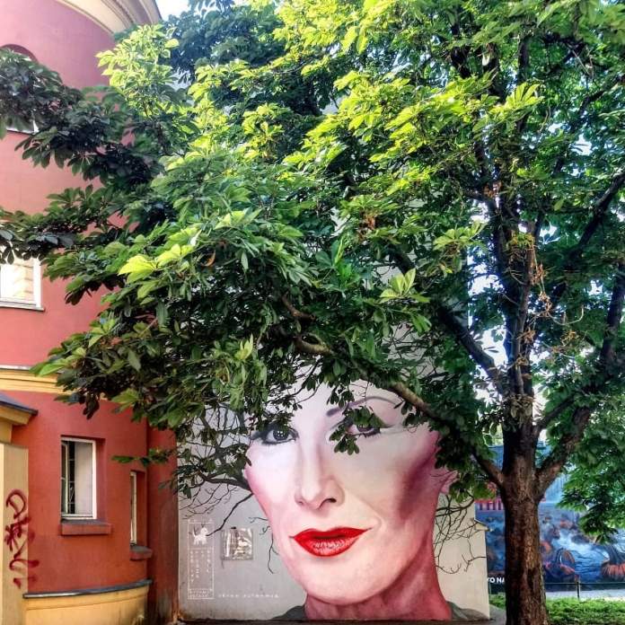 Summer view of the Kora mural by Bruno Althamer in Warsaw, Poland. The mural shows Kora's face with a serene expression and vivid red lips. A large tree with lush green leaves partially obscures the mural, giving the illusion that the foliage forms Kora's hair. The mural is located on a red building, and the greenery brings life to the portrait.