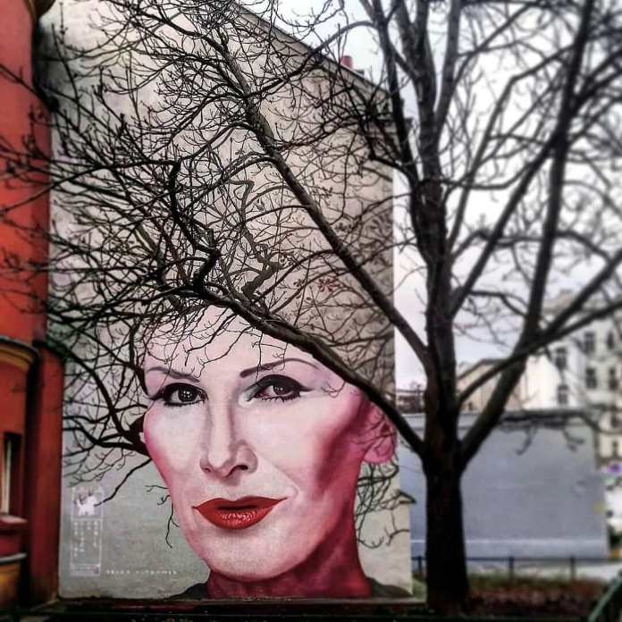Winter view of Bruno Althamer's mural in Warsaw, Poland, portraying Polish singer Kora. The tree in front of the mural is bare, with intricate branches creating an illusion of hair framing Kora's painted face. The mural's red lips and expressive eyes contrast against the subdued, wintry backdrop of the scene.