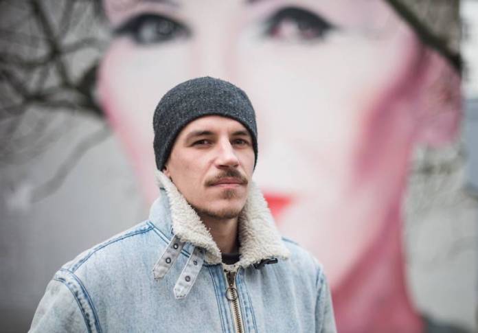 A portrait of street artist Bruno Althamer standing in front of his mural of Polish singer Kora (Olga Jackowska) in Warsaw, Poland. Althamer is wearing a black beanie and a denim jacket with a shearling collar, looking confidently at the camera. The mural, featuring Kora's face with red lips and expressive eyes, serves as the background, blending seamlessly with the tree's branches.