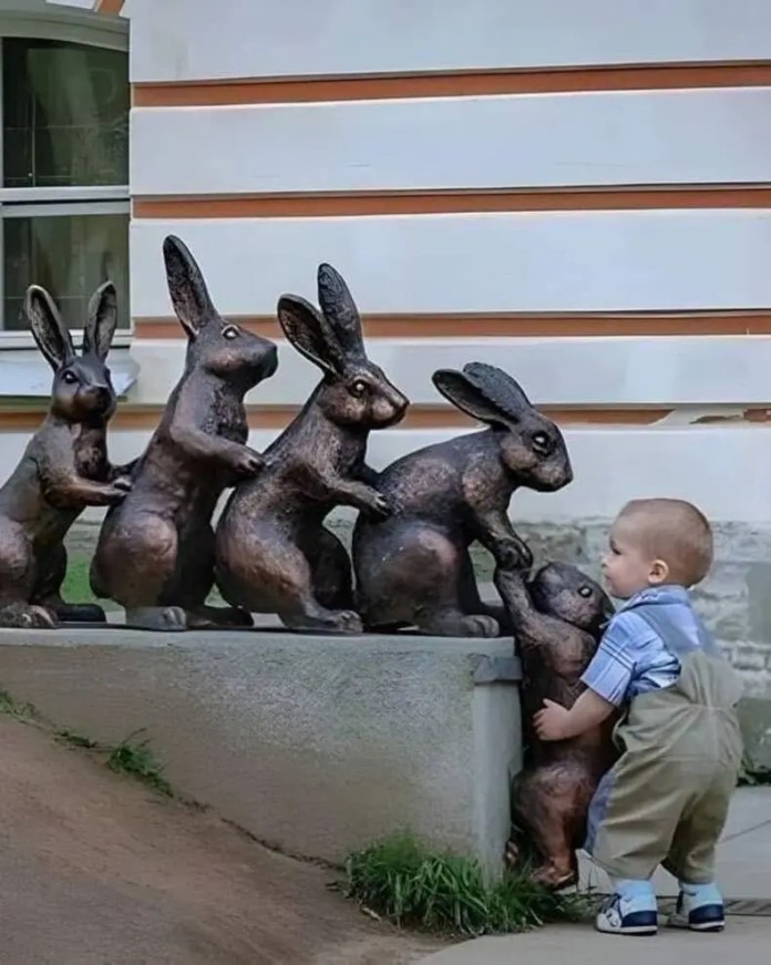 A toddler in overalls stands at the end of a line of bronze rabbit statues, mimicking their upright posture. The sculpture is set against a striped building wall.