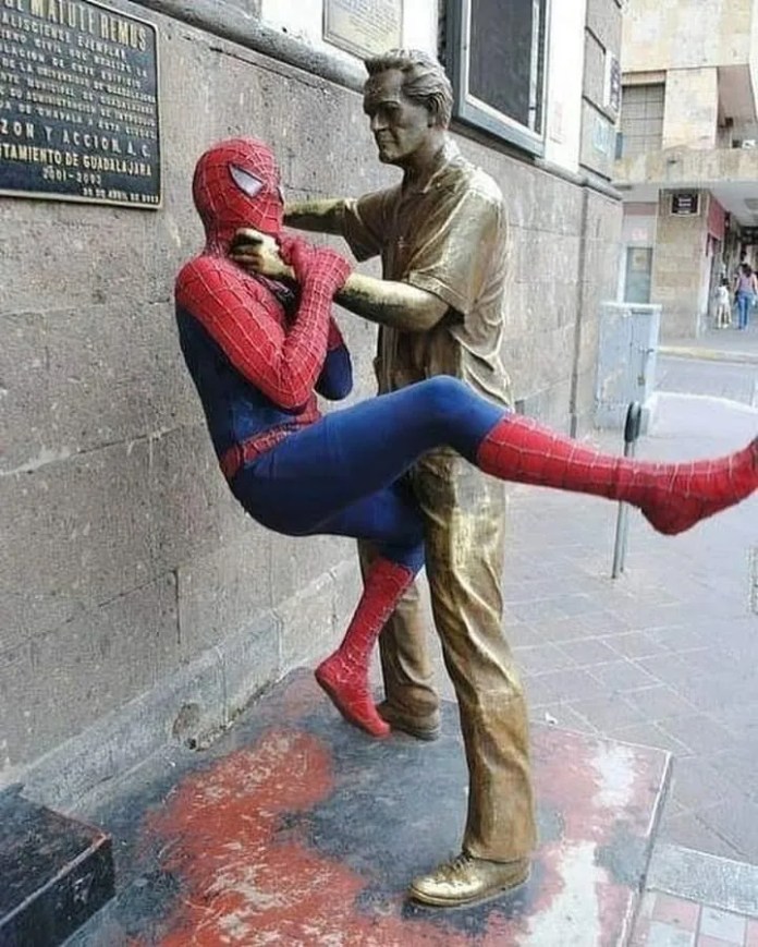 A person dressed as Spider-Man is lifted by the collar against a wall by a bronze statue of a man in an alley, creating a comic-book-style scene.