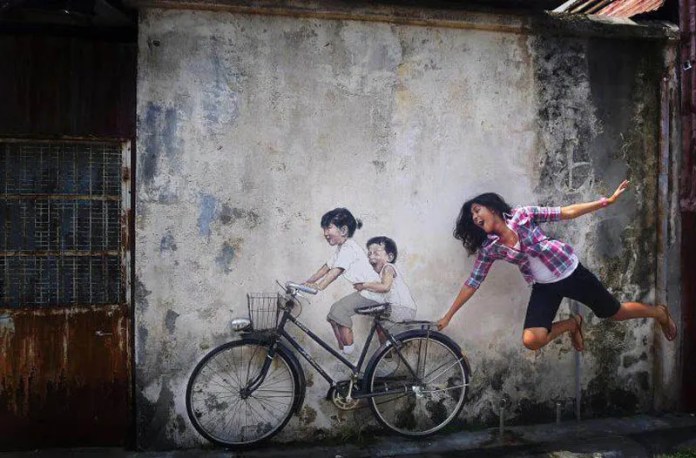 Mural in George Town, Malaysia by Ernest Zacharevic showing two children riding a real bike with painted bodies and faces, while a woman jumps behind them pretending to be pulled.