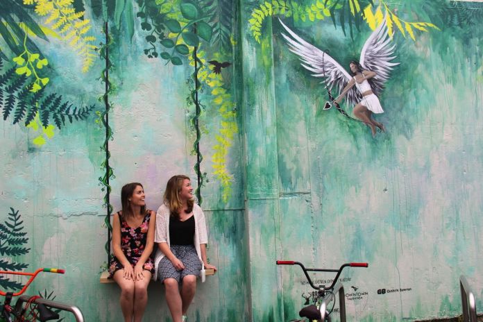 Interactive mural in downtown Tauranga, New Zealand by Millie Newitt and Tara Fowler, with real swings in front of painted jungle vines and a winged cupid about to shoot an arrow.