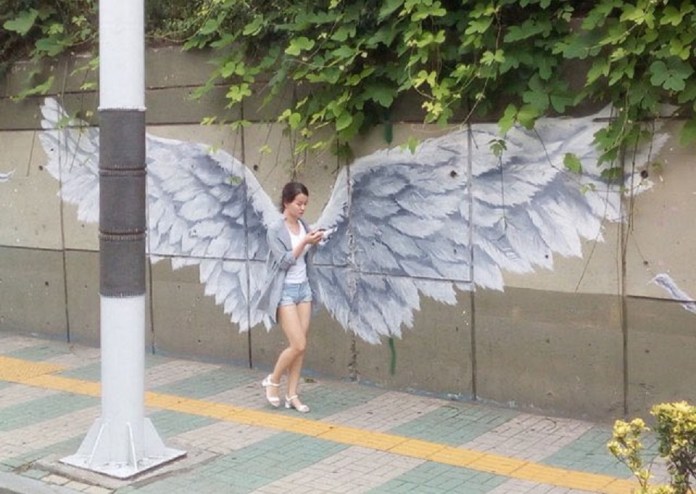 A pedestrian walks in front of a mural on a concrete wall in South Korea featuring large white angel wings, giving the illusion that the wings belong to her.
