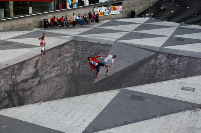 3D illusion artwork titled “Mind Your Step” by Erik Johansson at Plattan in Stockholm, Sweden, showing a giant pit in the pavement where people pose as if falling into a void.