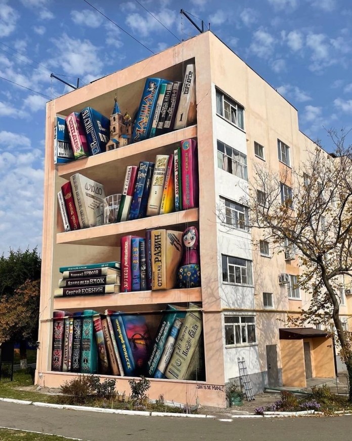Large mural on the side of an apartment block in Solnechnodolsk, Russia, painted to resemble four giant bookshelves filled with oversized books, a Matryoshka doll, and miniature props.