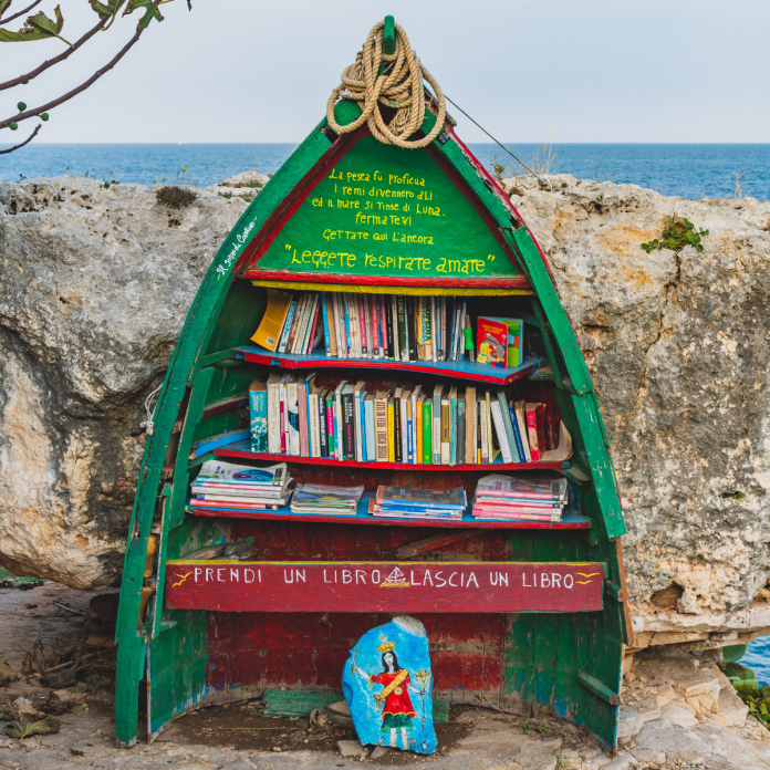 A colorful old wooden boat repurposed as a public bookshelf, filled with various books and placed beside a rocky seashore in Puglia, Italy. Painted messages in Italian encourage reading and community sharing.