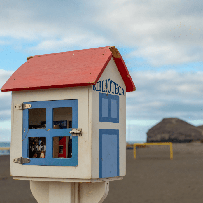 A tiny house-shaped public library painted white, red, and blue, sitting on a beach in Tenerife with volcanic mountains in the distance and books visible through its glass front.