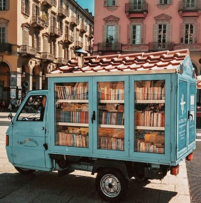 A vintage three-wheeled truck converted into a mobile library with sky-blue panels and a ceramic-tiled roof, filled with rows of books, parked in a historic Italian square.