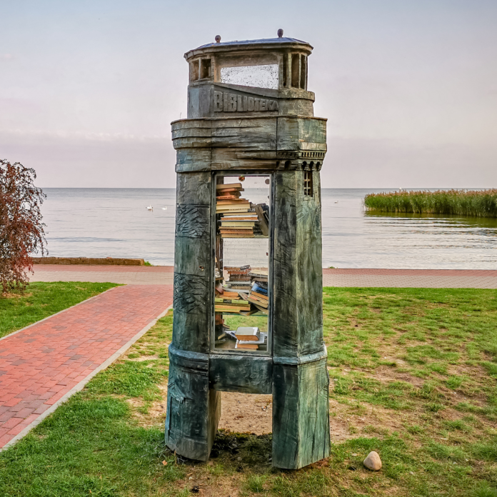 A tall lighthouse-shaped public bookshelf with a metallic, aged surface, placed near a lakeside walking path in Poland, its shelves stocked with various books.
