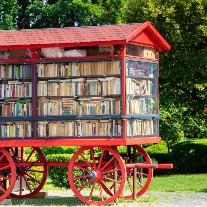 A large, old-fashioned red wagon turned into a bookmobile, fully stocked with neatly arranged books, situated in a green public park with trees in the background.