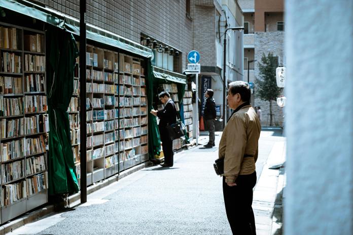 A narrow alley lined with tall bookshelves packed with Japanese books, partially shaded by green awnings, as a few people quietly browse the titles in Tokyo’s historic book district.
