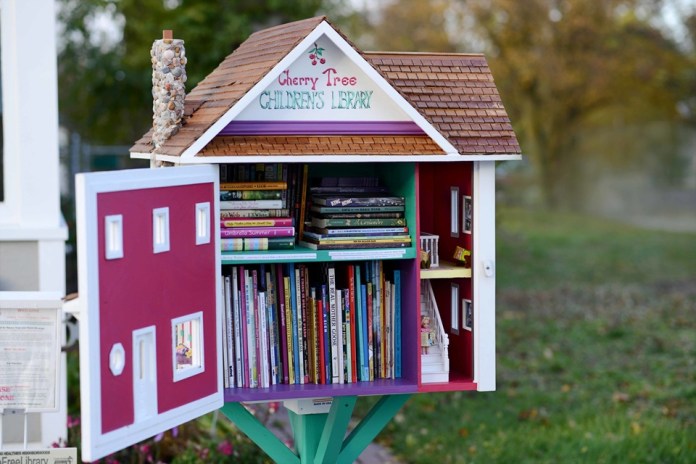 A small, house-shaped public library painted in pastel colors with intricate detail, stocked with children’s books and doll-sized furniture, designed like a miniature two-story house.