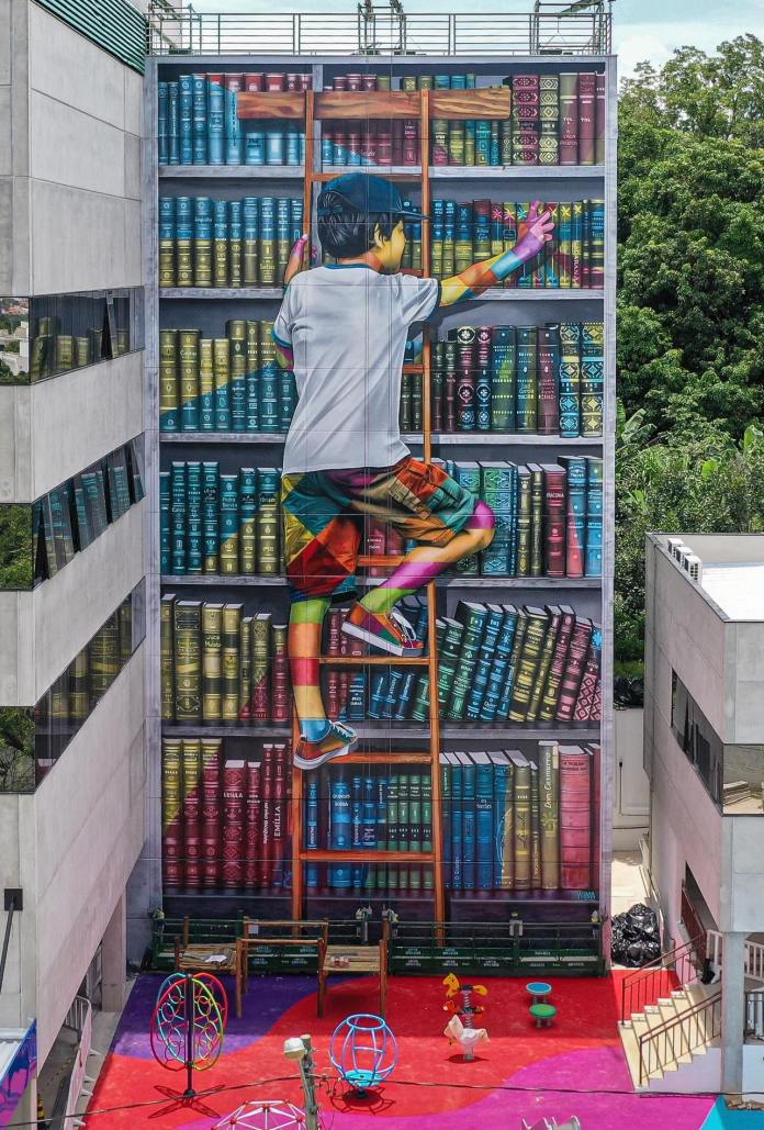 Mural by Eduardo Kobra in Sorocaba, Brazil, depicting a young boy climbing a wooden ladder on a massive painted bookshelf filled with colorful book spines across the side of a tall building.