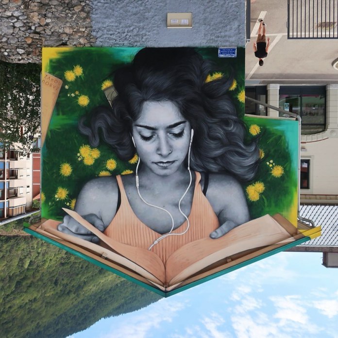 Mural by Zabou in Moutiers, France, showing a young woman reading a book while lying in green grass with yellow dandelions. The artwork is painted upside-down on the side of a building.