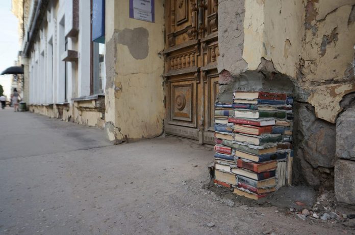 Urban art in Yekaterinburg, Russia, showing a damaged wall corner rebuilt using stacked real books embedded in cement, merging literature and architecture.