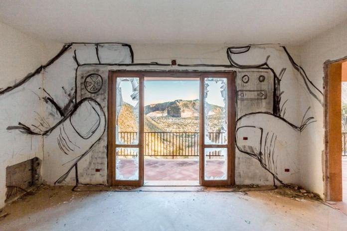 Mural of two hands holding a camera drawn around a doorway, with the landscape outside framed like a photo, painted inside a decaying room.