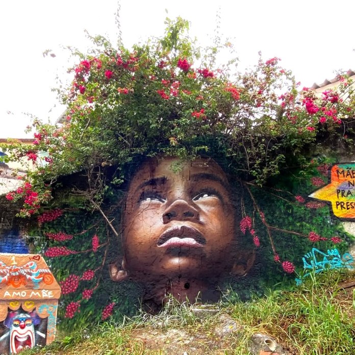Mural of a child’s face looking upward with expression of wonder, integrated with real overhanging flowers that appear as hair, painted on a garden wall.