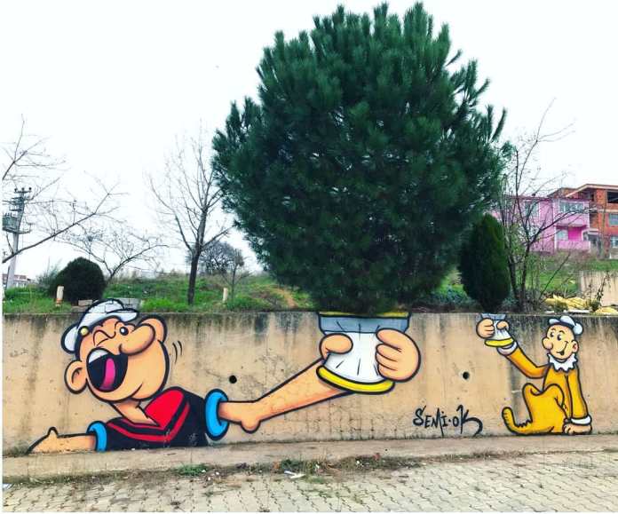 Street mural of Popeye handing a spinach can to another character, with a real tree positioned above the can to mimic spinach bursting out, painted along a low wall in Istanbul.